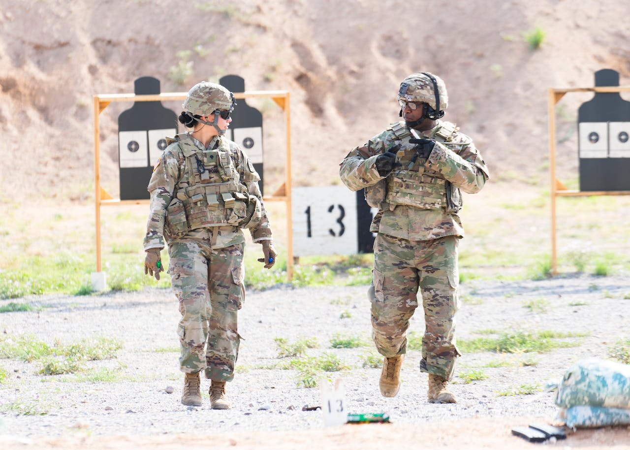 Two soldiers in military uniforms practicing at an outdoor shooting range with targets visible.